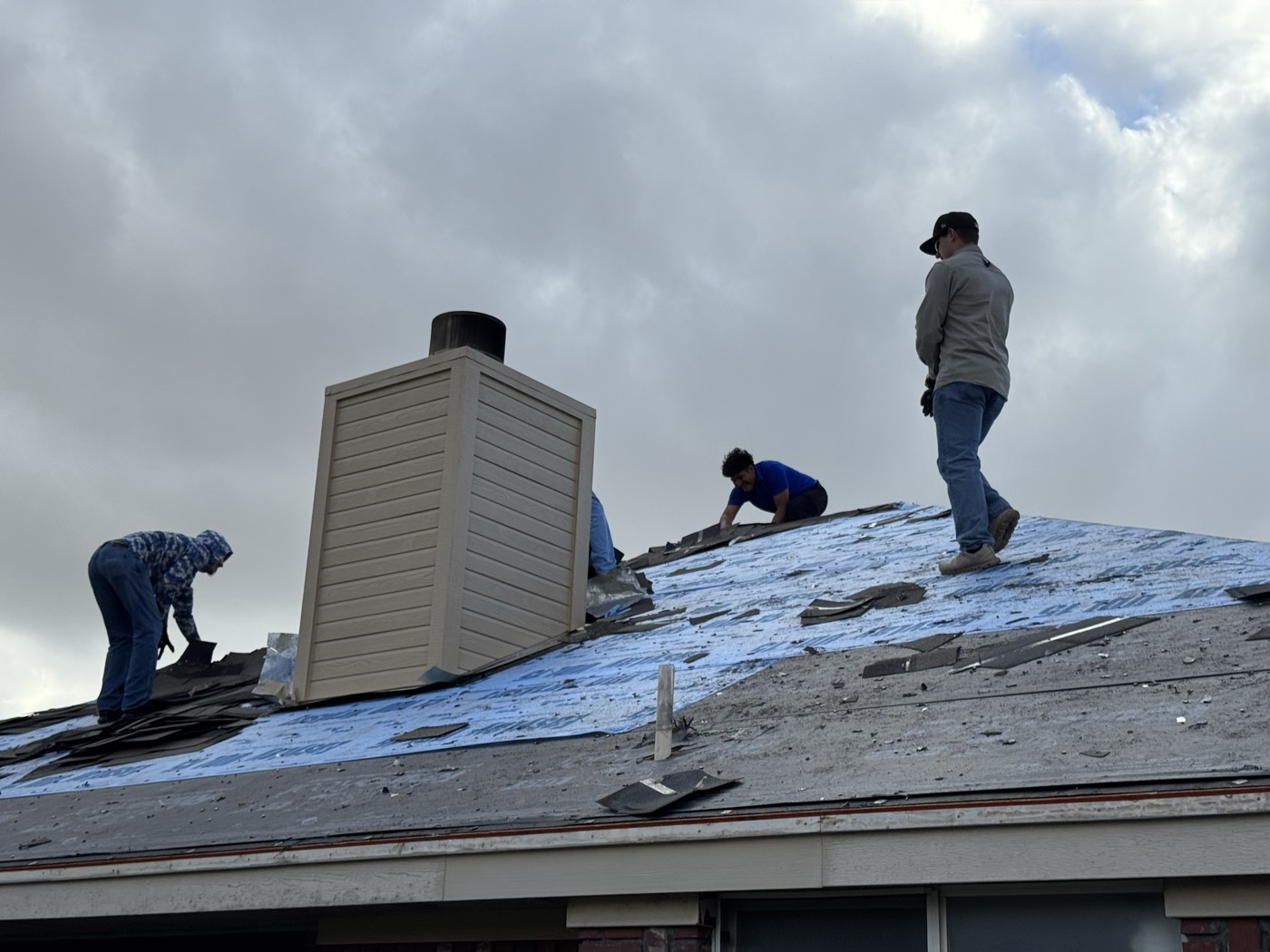 Three roofers stripping shingles around a rebuilt chimney chase under a heavy gray sky in Leander, TX